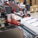 Nahaufnahme einer Pharma Serialisierungslinie bei der MSK: Ein hochpräziser Drucker bringt einen Matrix-Code, die GTIN, Serialnummer und das Verfallsdatum auf eine Medikamentenfaltung auf.